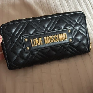 Svart quiltad plånbok från Love Moschino - Snygg svart plånbok från Love Moschino med quiltad design och guldfärgade detaljer. Plånboken har dragkedja runtom, flera fack för kort och mynt samt en lyxig känsla med skinnmaterial. Perfekt för dig som vill ha både stil och funktion. Väskan är som ny den är nästan aldrig använd och den är ÄKTA.
