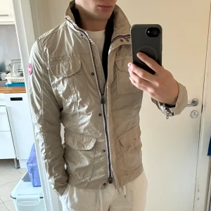 Beige fältjacka från Colmar - Snygg beige fältjacka från Colmar med hög krage och dragkedja framtill. Jackan har flera fickor med lock och tryckknappar, samt Colmar-logga på ärmen. Tillverkad i lätt och glansig polyester, perfekt för vår och höst. Bra skick, lite smuts på ena fickan. Har modifierats för bättre form på kemtvätt.