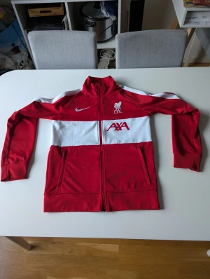 Liverpool Nike ziptröja röd/vit - Säljer en snygg röd och vit ziptröja från Nike med Liverpool FC-logga och AXA-tryck på bröstet. Tröjan har hel dragkedja, ståkrage och långa ärmar. Perfekt för dig som älskar fotboll och vill visa ditt stöd för LFC.