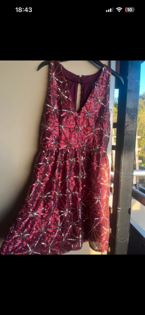 Glittrig vinröd kortklänning julig - Säljer en glittrig vinröd kortklänning med silverpaljetter i geometriskt mönster. Klänningen har ärmlös design och en snygg v-ringning med knapp i nacken. Perfekt för dig som vill sticka ut med lite extra bling. Prislappen sitter kvar