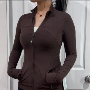 Brun sportig zipjacka med fickor - Slimmad brun zipjacka från Lululemon med hög krage och långa ärmar. Jackan har praktiska fickor med dragkedja och snygga sömdetaljer som framhäver siluetten. Perfekt för dig som gillar en sportig och clean look.