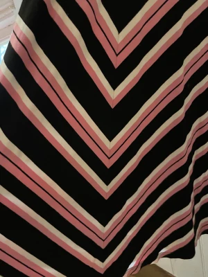 Randig långklänning från Jumperfabriken - Supersnygg långklänning från Jumperfabriken i svart, rosa och vit med coola diagonala och horisontella ränder. Klänningen har omlott-v-ringning och trekvartsärm. Mjuk och stretchig polyester som sitter skönt och ger en riktigt chic look.