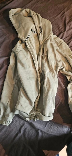 Grön dragkedje hoodie från Sail Racing - Snygg grön hoodie från Sail Racing med huva och dragkedja framtill. Hoodien har en bröstficka med dragkedja och mjukt teddyfoder på insidan. Perfekt för kyliga dagar och har en clean look med diskret logga på bröstet.