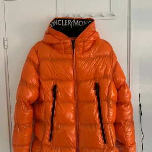 Orange Moncler pufferjacka med huva - Säljer en fet orange pufferjacka från Moncler med stor huva och svarta detaljer. Jackan har två långa dragkedjor framtill, Moncler-logga på ärmen och coolt tryck på insidan. Perfekt för dig som vill sticka ut i vinter och hålla dig varm. Pris kan diskuteras.