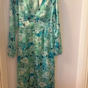 Blommig långklänning från Adoore - Säljer en superfin långklänning från Adoore med blommigt mönster i turkosa, gröna och vita toner. Klänningen har v-ringning, långa ärmar och knappar framtill. Materialet är mjukt och lätt, perfekt för vår och sommar. Snygg passform och flowy känsla.
