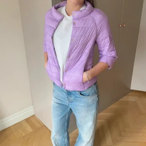 Ljuslila quiltad pufferjacka - Säljer en ljuslila pufferjacka med quiltad design och guldfärgade knappar framtill. Jackan har en rundad krage och trekvartsärmar, vilket ger en chill och modern vibe. Perfekt att slänga på sig när det är lite kyligt ute.