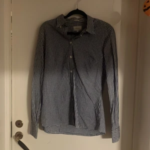 Blårutig skjorta från Boomerang - Snygg blårutig skjorta från Boomerang i trim fit-modell. Skjortan har klassisk krage, långa ärmar med manschett och knappar framtill. Tillverkad i mjuk bomull som känns skön mot huden. Perfekt för dig som gillar stilrena och tidlösa mönster.