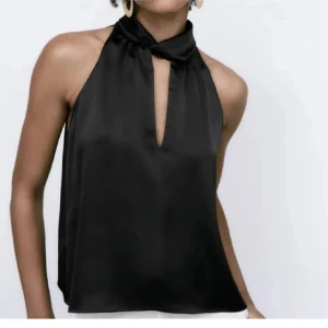 Svart halterneck festtopp i satin - Säljer en svart halterneck topp i glansigt satinmaterial med snygg knytning i halsen och djup v-ringning framtill. Använt ett fåtal ggr. 