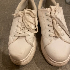 Vita sneakers med platåsula - Säljer ett par vita sneakers med platåsula och snörning. Skorna har en klassisk rund tå och är tillverkade i syntetmaterial. Baktill finns en svart detalj som ger en snygg kontrast. Perfekta för dig som gillar en clean och stilren look.