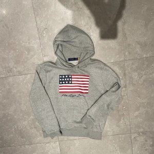 Grå Ralph Lauren Hoodie - Snygg grå hoodie från Polo Ralph Lauren i storlek M. Tröjan har en stor amerikansk flagga tryckt på bröstet och Polo Ralph Lauren-logga under. Mjuk bomullsmix, huva med snörning och ribbade muddar. Perfekt för chill dagar och streetstyle.