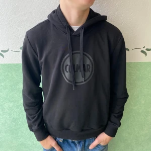 Svart Colmar hoodie med logga - Säljer en svart hoodie från Colmar i storlek M. Tröjan har en stor Colmar-logga på bröstet, snörning i huvan och ribbade muddar vid ärmslut och nederkant. Perfekt för dig som gillar stilrena och bekväma plagg.