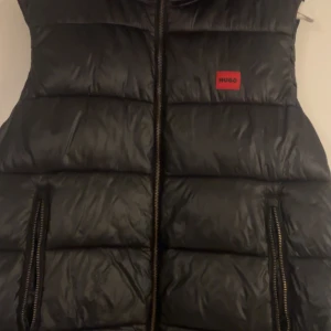 Svart dunväst från Hugo med huva - Svart dunväst från Hugo med röd logotyp på bröstet. Västen har huva, två fickor med dragkedja och quiltad design. Materialet är glansigt och känns lätt. Perfekt för lager-på-lager och streetstyle. Dragkedja framtill och snygga detaljer. DEN HAR EN SKAVANK PÅ DRAGKEDJAN SÅ DRT ÄR DARFÖR JAG SÄLJER TILL SÅ BILLIGT