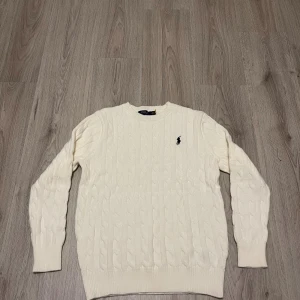 Vit kabelstickad tröja Polo Ralph Lauren - Kabelstickad tröja från Polo Ralph Lauren i mjuk vit bomull. Klassisk rund hals och långärmad modell med ribbade muddar. Ikonisk broderad logga i svart på bröstet. Perfekt för dig som gillar clean och stilren look.