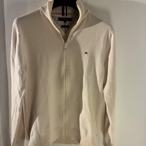 Tommy Hilfiger – The 1985 Sweater (stl M) - Säljer en cremevit zip-tröja från Tommy Hilfiger, modell The 1985 Sweater, i storlek M. Tröjan är i utmärkt skick och väl omhändertagen. Som du kan se på bilderna är den inte strykt, vilket gör att vissa veck syns, men plagget är i fint skick i verkligheten. Den har hög krage med Hilfigers randdetalj, dragkedja framtill och ett litet broderat logomärke på bröstet. Nypris brukar ligga runt 1 199–1 499 kr.