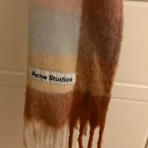 Rutig halsduk från Acne Studios - Supermjuk och fluffig halsduk från Acne Studios med rutigt mönster i ljusa toner av beige, brunt, rosa och blått. Klassisk fransad kant och stor logotyp-patch. Perfekt accessoar för att lyfta din vinterstil. Mycket bra skick, endast använd en vinter. Pris går att diskutera!!💓