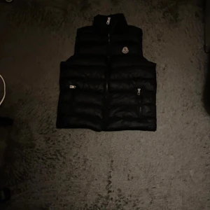Svart dunväst - Svart dunväst med quiltad design och hög krage. Västen har två fickor med dragkedja framtill och Moncler-logga på bröstet. Insidan har ett unikt serietidningsmärke. Perfekt för lager-på-lager under kyliga dagar.