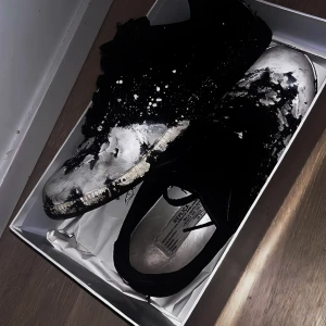 Maison Margiela gats - Maison Margiela sneakers i svart mocka med unikt vitt färgstänksmönster över hela skon. Låg modell med snörning och rund tå. Snygg kontrast mellan svart och vitt, perfekt för dig som gillar statement pieces och streetstyle.