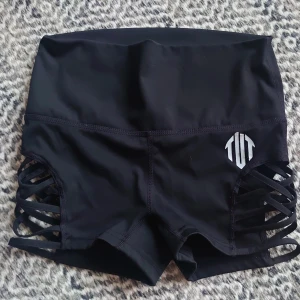 Svarta shorts med sidoband från Tut Empire  - Snygga svarta shorts från TUT Empire med hög midja och coola öppna sidor med korsade band. Tillverkade i stretchigt material med 88% nylon och 12% spandex, perfekta för träning eller dans. Logga i vitt framtill ger extra attityd.