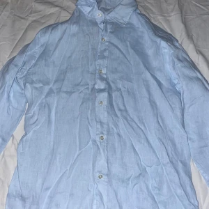 Oscar Jacobson Skjorta - Säljer en stilren ljusblå skjorta i linne med klassisk krage och knäppning framtill. Skjortan har lång ärm och en luftig känsla, perfekt för varmare dagar. Enkel och clean look som passar till många outfits.