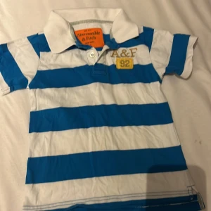 Randig pikéskjorta Abercrombie & Fitch - Snygg pikéskjorta från Abercrombie & Fitch i storlek S baby storlek. Skjortan har breda blå och vita ränder, klassisk krage och korta ärmar. På bröstet finns broderad A&F-logga och en gul patch med siffran 92. Perfekt för en chill och sportig look.