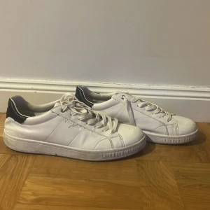 Vita sneakers från Björn Borg strl 44 - Snygga vita sneakers från Björn Borg med svart detalj på hälen. Klassisk låg modell med rund tå och platt sula. Skorna har snörning och är tillverkade i skinn för en clean och stilren look. Perfekta för dig som gillar minimalistisk stil.