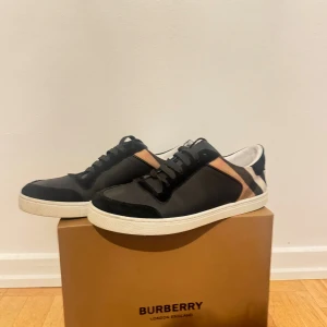 Burberry sneakers med rutigt mönster - Snygga svarta sneakers från Burberry med klassiskt rutigt mönster i beige, svart och vitt på sidorna. Skorna har svarta snören, vit platt sula och detaljer i skinn och mocka. Perfekta för dig som vill ha en stilren men ändå unik look.