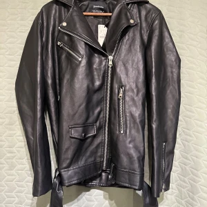 Oversize bikerjacka i skinneffekt - Svart oversize bikerjacka/skinnjacka från Stradivarius i damstorlek S. Detalj med hällor och tryckknappar på axlarna. Bälte nedtill med spänne. Köpt för 559kr. ❕Helt ny. Aldrig använd. Prislapp kvar.❕ Längd: ca 70cm, Bredd: ca 52cm
