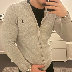 Grå kofta från Ralph Lauren - Snygg grå kofta från Ralph Lauren med dragkedja framtill och klassisk svart logga på bröstet. Tröjan har ribbade muddar och krage, långärmad och tillverkad i mjuk bomull. Perfekt för dig som gillar stilrena och bekväma plagg.