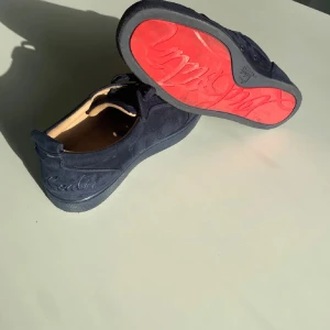 Christian Louboutin mörkblå sneakers - Snygga mörkblå sneakers från Christian Louboutin med klassisk röd sula och broderad logga på hälen. Skorna är gjorda i mjuk mocka med rund tå, snörning och beige skinnfoder. Perfekt för dig som vill sticka ut med exklusiva detaljer och lyxig känsla.