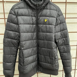 Svart pufferjacka från Lyle & Scott - Svart pufferjacka från Lyle & Scott med gul logotyp på bröstet. Jackan har huva, ribbade muddar och dragkedja framtill. Quiltad design med horisontella sömmar och två sidofickor. Perfekt för kyliga dagar och har en clean, sportig look. Litet märke på ena ärmen