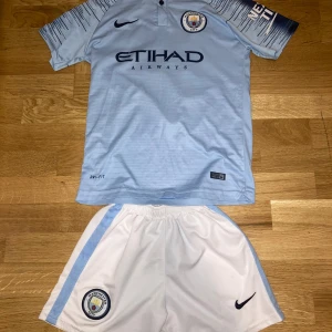 Manchester City De Bruyne fotbollströja - Manchester City matchtröja med De Bruyne 17 på ryggen, tillverkad i Dri-Fit material från Nike. Tröjan är ljusblå med mörkblå detaljer och korta ärmar, har klubbmärke och sponsortryck på bröstet samt mönstrade ärmar. Tillhörande vita shorts med ljusblå ränder och klubbmärke.