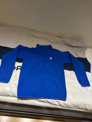 Blå vindjacka från Adidas - Helt oanvänd Snygg blå vindjacka från Adidas med klassisk logga på bröstet. Jackan har hel dragkedja, ståkrage och långa ärmar. Perfekt för dig som gillar sportig stil och vill ha något lätt och bekvämt. Materialet är polyester och jackan har en clean look utan mönster.