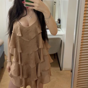 Beige volangklänning💕 - Säljer en superfin beige kortklänning med flera lager volanger och omlott-design. Klänningen är ärmlös och har en djup v-ringning, perfekt för dig som gillar en flowy och feminin stil. Materialet känns lätt och luftigt, perfekt för varma dagar.
