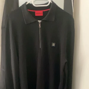 Hugo boss cardigan - Svart pikétröja från Hugo med klassisk krage och half zip framtill. Tröjan har lång ärm och en diskret Hugo boss logga på bröstet.