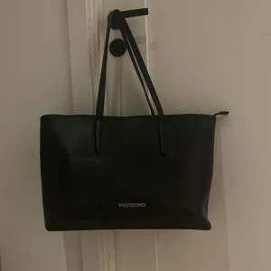 Svart shoppingväska från Valentino - Stilren svart shoppingväska från Valentino i slätt skinn med guldiga detaljer och dragkedja upptill. Väskan har långa handtag och rymligt innerfack med extra ficka. Perfekt för dig som vill ha en klassisk och snygg väska med plats för allt du behöver.