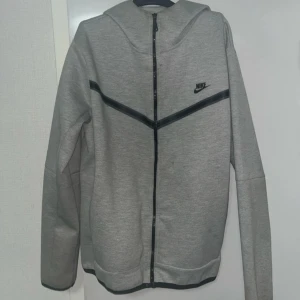 Grå hoodie från Nike med dragkedja - Grå hoodie från Nike i storlek L med hel dragkedja och svart Nike-logga på bröstet. Tröjan har huva, långa ärmar och svarta detaljer i form av ett V-format band över bröstet. Mjuk och skön, perfekt för chill eller träning.