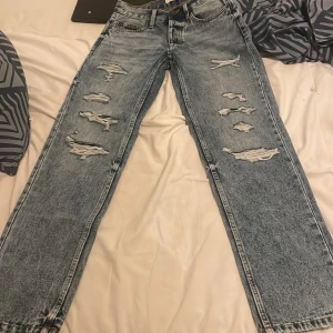 Distressed jeans från FSBN, blå 27/32 - Säljer ett par blå jeans från FSBN i storlek 27/32 med snygga slitningar och trasiga detaljer framtill. Jeansen har rak passform, klassisk femficksdesign och är tillverkade i jeansmaterial. Perfekt för dig som gillar en edgy och avslappnad stil.