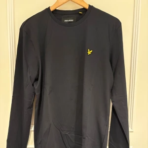 Långärmad tröja Lyle & Scott - Svart långärmad tröja från Lyle & Scott med klassisk rund hals och gul broderad logga på bröstet. Tröjan är gjord i mjuk bomull och har en clean, stilren look som funkar till jeans eller chinos. Perfekt för dig som gillar enkel och snygg stil. Tröjan är i storlek M och är helt ny och oanvänd