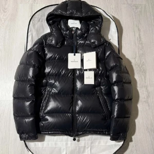 Svart Moncler pufferjacka med huva - Svart pufferjacka från Moncler med glansig finish, huva och dragkedja framtill. Jackan har Moncler-logga på ärmen, två sidofickor med dragkedja och quiltad design. Perfekt för kalla dagar och har en modern, clean look som sticker ut.