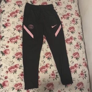 Svarta PSG träningsbyxor från Nike - Svarta träningsbyxor från Nike med rosa detaljer och PSG-logga på ena benet. Slimmad passform med hög midja och smala ben. Perfekta för fotboll eller träning, tillverkade i mjukt syntetmaterial som andas. Snygg design med rosa Nike-swoosh och paneler. Pris kan även diskuteras.