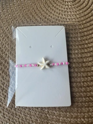 Pärlarmband med sjöstjärna - Sött hemmagjord armband med små rosa och vita pärlor och en vit sjöstjärna i mitten. Perfekt för dig som gillar strandvibbar och färgglada detaljer. Armbandet är enkelt och har en lekfull design som sticker ut.