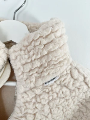 Beige teddyjacka från Acne Studios - Säljer en beige teddyjacka från Acne Studios med halv dragkedja och hög krage. Jackan har en fluffig, krusig yta i teddy-material och insida i bomull. Modellen är loose fit med stora fickor och långärmad design. Perfekt för kalla dagar och riktigt snygg till jeans.