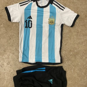 Argentina Messi fotbollströja Adidas - Säljer en klassisk Argentina fotbollströja från Adidas med Messi och nummer 10 på ryggen. Tröjan har blå och vita vertikala ränder, svarta detaljer och korta ärmar. Materialet är lätt och andas, perfekt för match eller träning. AFA-emblem på bröstet.