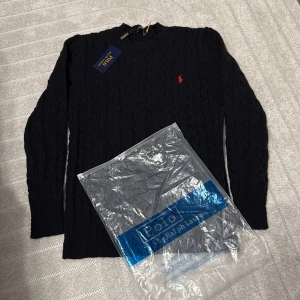Svart kabelstickad tröja Polo Ralph Lauren - Kabelstickad svart tröja från Polo Ralph Lauren med klassisk röd broderad logga på bröstet. Tröjan har rund halsringning och långa ärmar, tillverkad i mjukt stickat material som ger en clean och tidlös look.