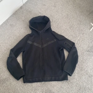 Svart Nike vindjacka med huva XL - Svart vindjacka från Nike i storlek XL med huva och hel dragkedja framtill. Jackan har diskreta grå detaljer över bröstet och en liten ficka med dragkedja på ena ärmen. Perfekt för dig som gillar sportig och clean stil.