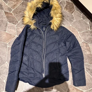 Mörkblå dunjacka med fuskpäls från Svea - Snygg mörkblå dunjacka från Svea med fluffig fuskpäls på huvan. Jackan har quiltad design, dragkedja framtill och långärmade ärmar. Perfekt för kalla dagar med sin varma och stilrena look. Svea-logga på bröstet och coolt mönster på fodret.