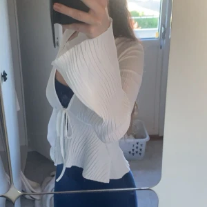 Vit omlottblus med snörning - Säljer en vit omlottblus med snörning framtill och vida, plisserade ärmar. Blusen har en lätt transparent look och vacker struktur i tyget som ger en cool vibe. Perfekt för dig som gillar trendiga detaljer och en luftig känsla.