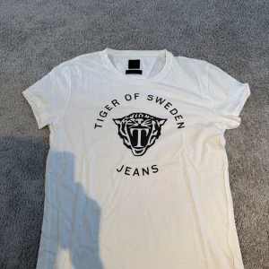 Vit Tiger of Sweden Jeans t-shirt - Vit t-shirt från Tiger of Sweden Jeans med svart tigertryck och logga på bröstet. Klassisk rund hals och korta ärmar. Materialet är mjuk bomull som känns skönt mot huden. Perfekt för dig som gillar stilrena och coola prints.