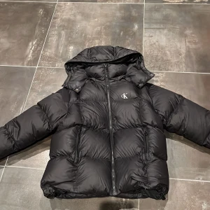 Pufferjacka från Calvin Klein - Svart pufferjacka från Calvin Klein med huva och dragkedja framtill. Jackan har CK-logga på bröstet och två sidofickor med dragkedja. Den är quiltad och har en loose passform, perfekt för kalla dagar. Materialet är syntet och dun, vilket gör den riktigt varm och skön. Det är ett litet röt märke som man kan se på bild 4 !! 
