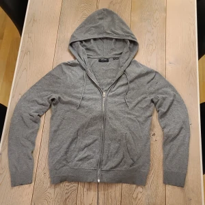 Grå stickad hoodie från Jack & Jones - Grå stickad hoodie från Jack & Jones i storlek S. Tröjan har huva med snörning, hel dragkedja framtill och två fickor. Materialet är mjukt och passformen är normal. Perfekt för chill dagar och enkel att matcha med jeans eller joggers.